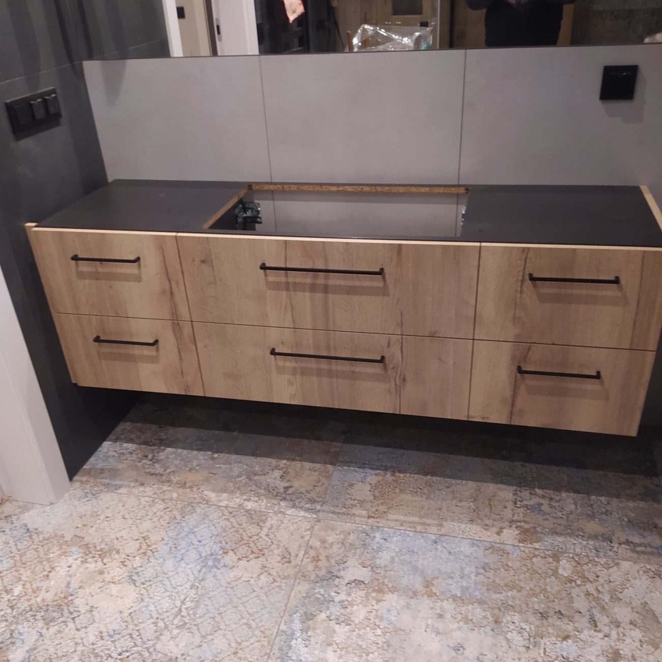 Łazienka z szarymi płytkami i drewnianą szafką pod umywalkę bez umywalki, na podłodze wielokolorowa mozaika